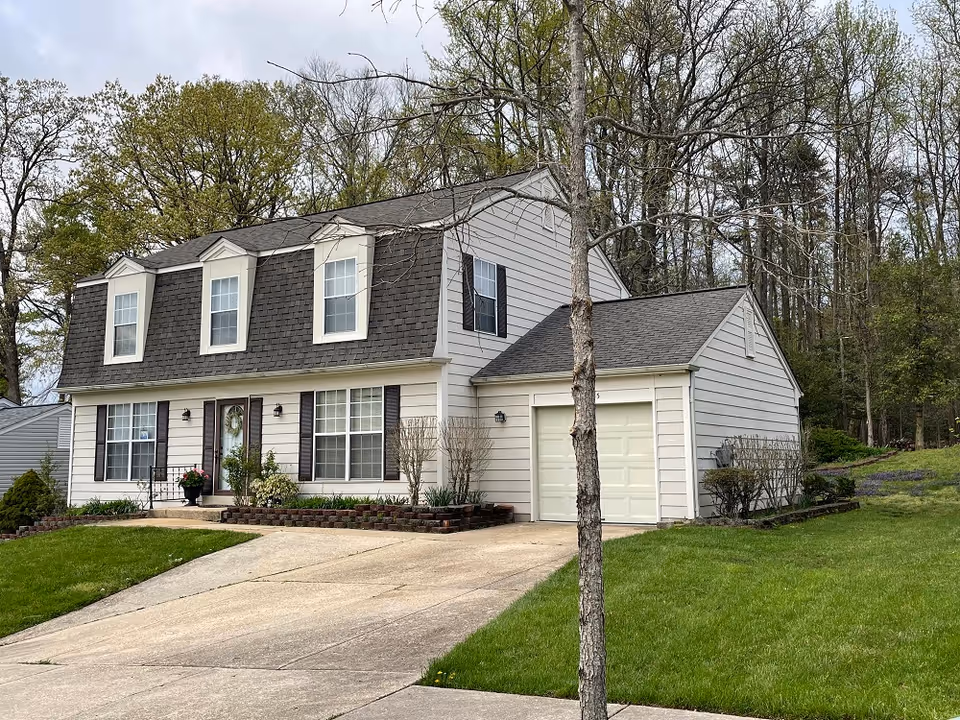 Front view of a two-story white-sided house with dormer windows, attached garage, driveway, and front lawn.