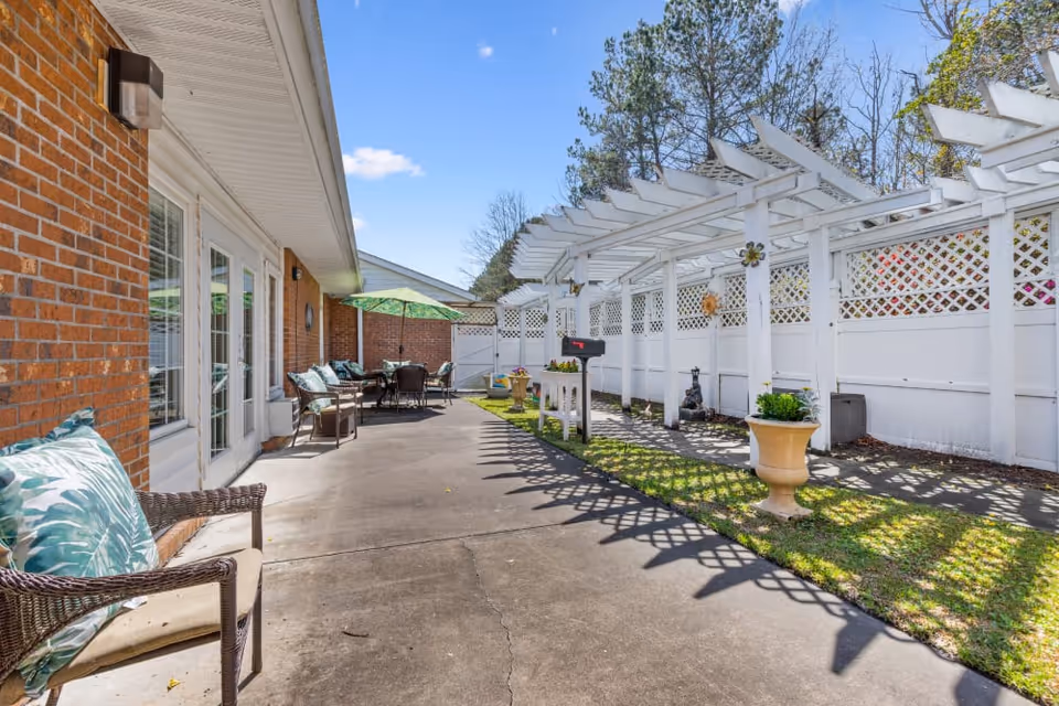 Sunny outdoor patio with wicker seating and a dining table beside a brick building and a white pergola over a walkway.
