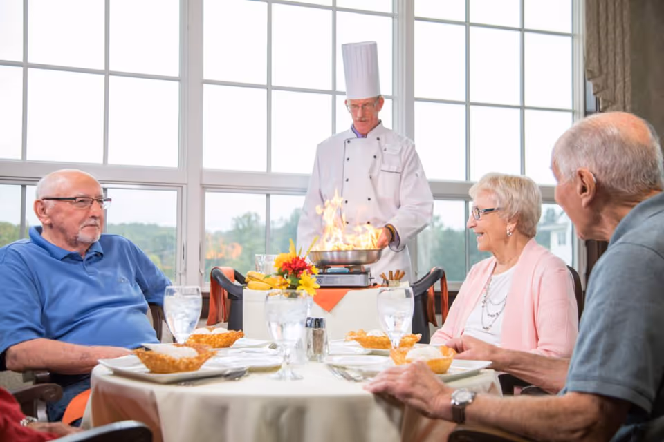 A chef flambéing a dish tableside while three elderly residents sit around a dining table set with water glasses and desserts.