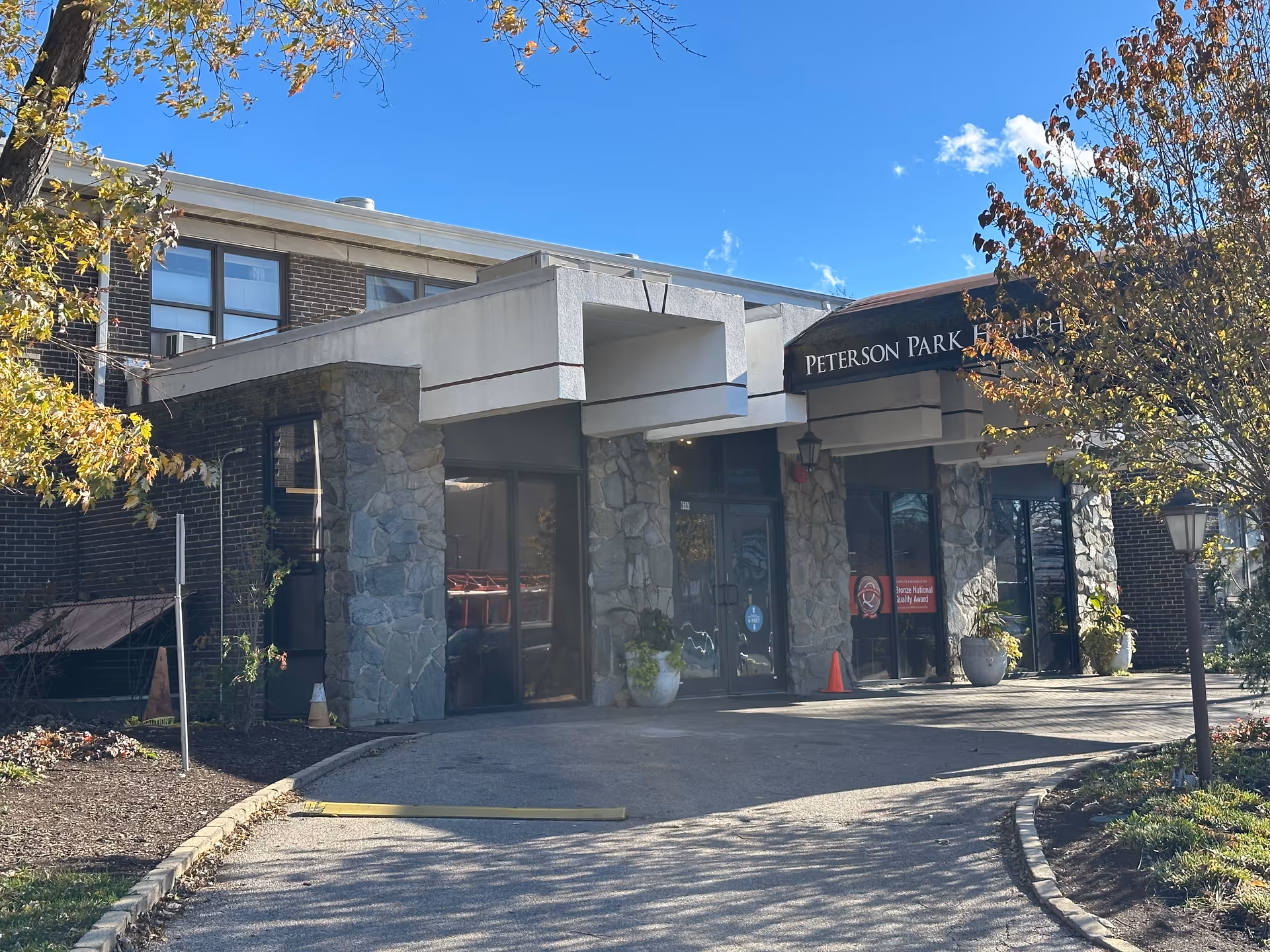 Front entrance of Peterson Park Health Care Center with a covered drop-off, stone pillars, and an awning under a clear blue sky.