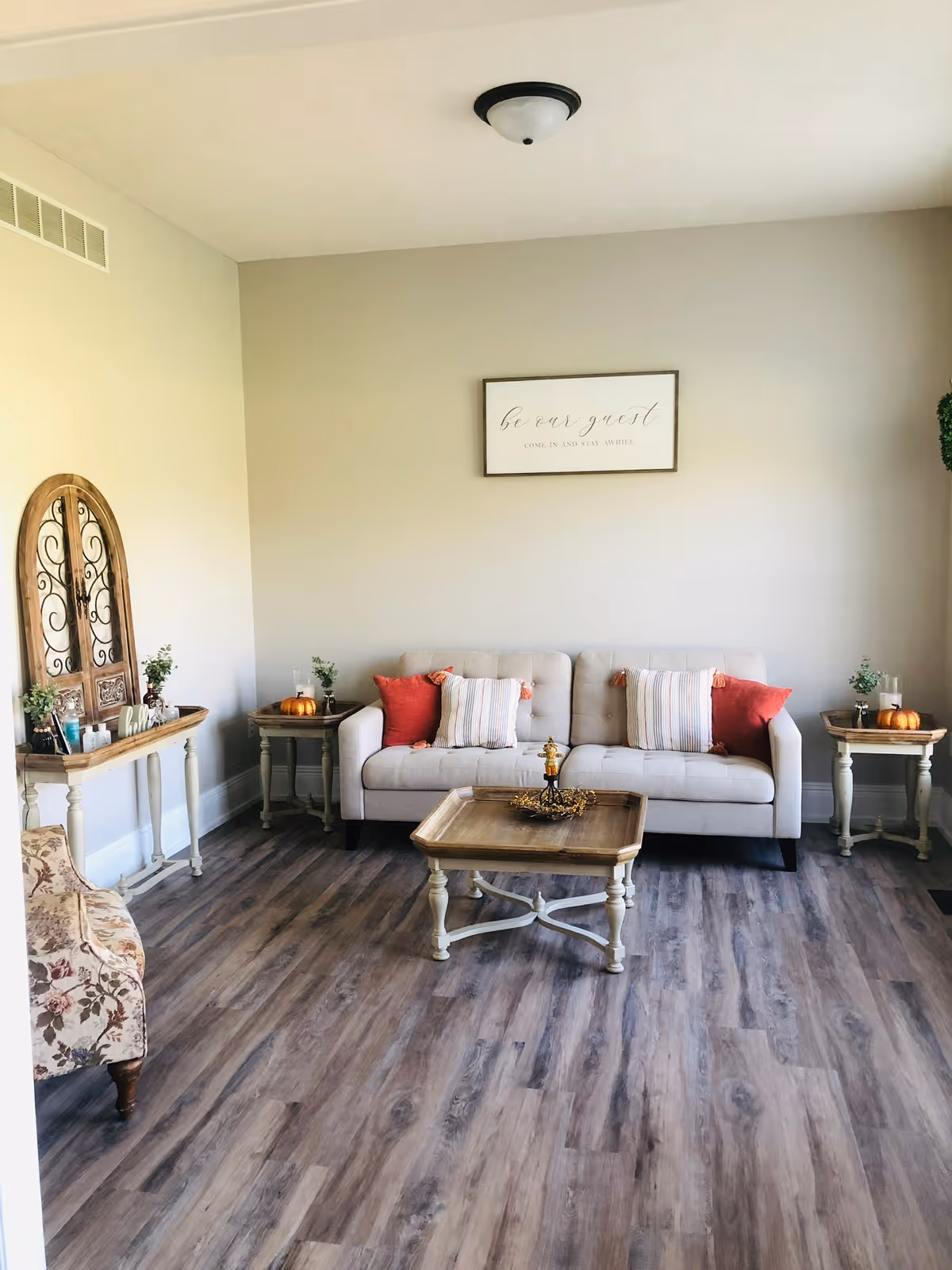 A cozy living room with a beige sofa adorned with red and striped pillows, a wooden coffee table in front, two matching side tables with small plants and decorative pumpkins, a floral armchair, and a wooden console table with decorative items. A framed sign on the wall reads 'be our guest, COME IN AND STAY AWHILE'. The room has wood flooring and neutral-colored walls.