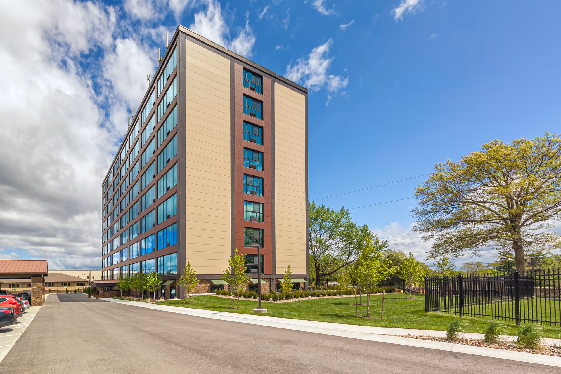 Multi-story residential building exterior with a landscaped lawn, trees, and adjacent parking under a partly cloudy blue sky.