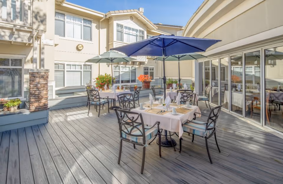 Outdoor patio area at Fremont Hills with several tables covered with light pink tablecloths, set for dining with plates, cups, and napkins. Each table has a large umbrella for shade, and there are potted plants around the perimeter. The patio is surrounded by the building's exterior walls with multiple windows and sliding glass doors.