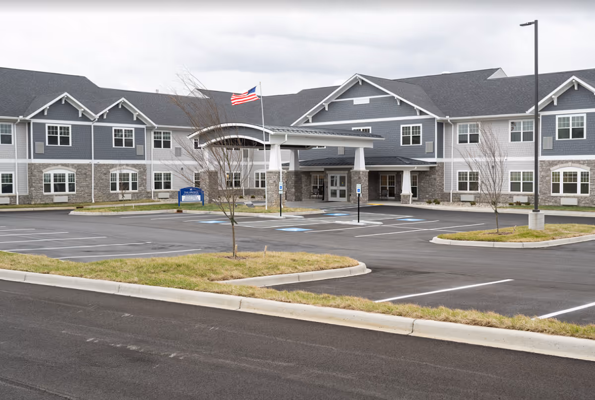 Exterior view of The Springs at Oldham Reserve senior living facility showing a large two-story building with gray and white siding, stone accents, multiple windows, a covered entrance with white pillars, an American flag on a flagpole, and an empty parking lot with marked handicap spaces.