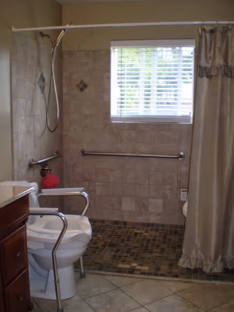 Bathroom with a walk-in shower featuring a handheld showerhead, grab bars on the tiled walls, a window with blinds, a shower curtain, and a toilet equipped with safety rails.