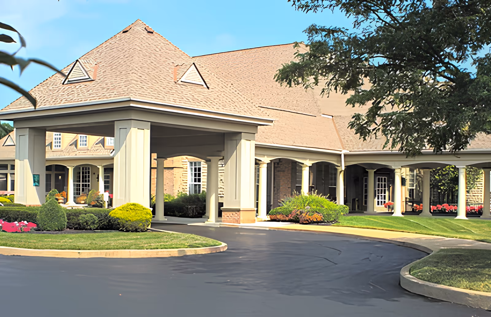 Exterior view of Feridean Commons facility showing a covered entrance with large pillars, a curved driveway, well-maintained landscaping with bushes and flowers, and a building with a shingled roof and multiple windows under a clear blue sky.