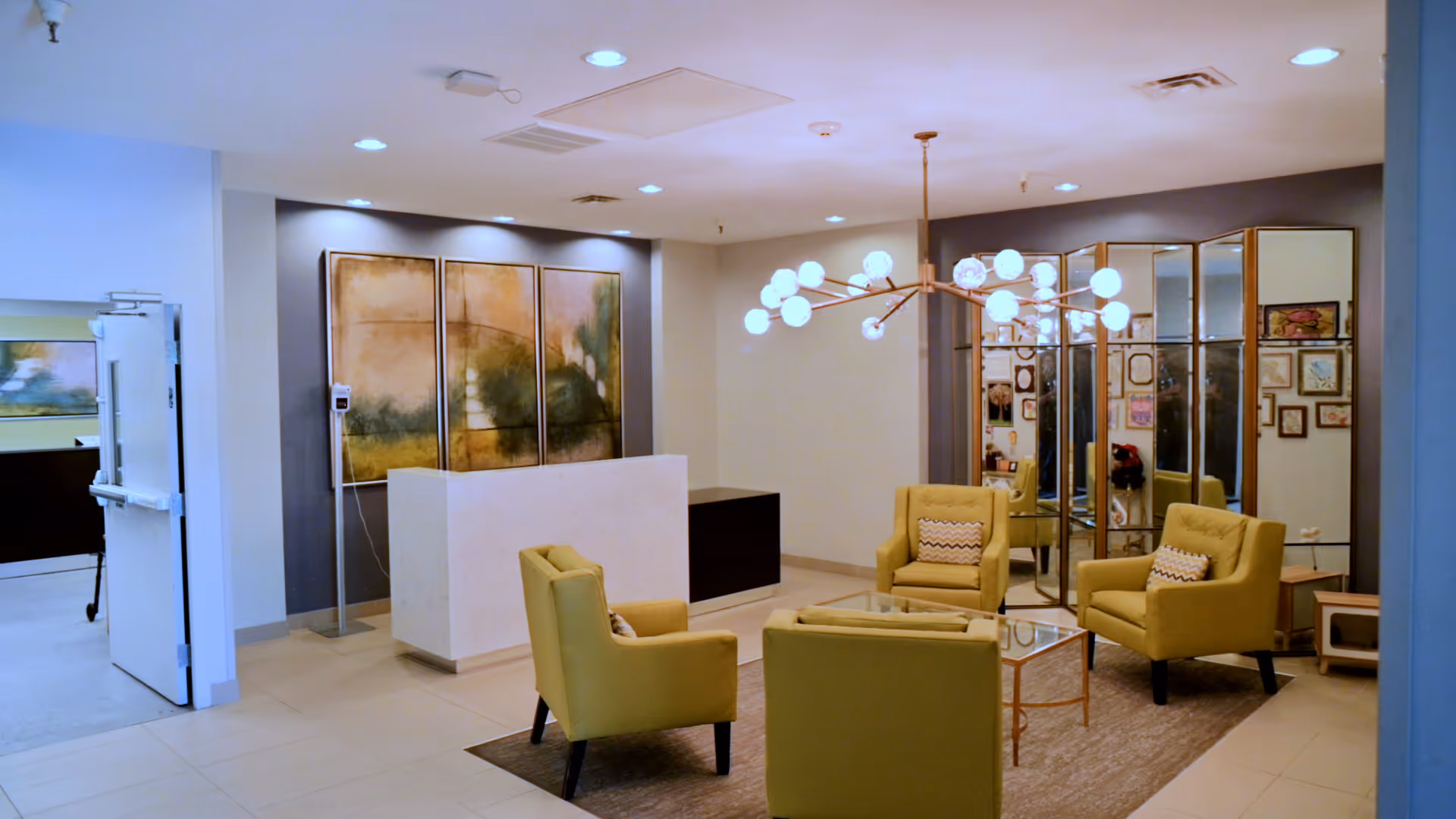 A modern and cozy seating area in a healthcare facility with four mustard yellow armchairs arranged around a glass coffee table on a rug. Behind the seating area is a decorative wall with large mirrors and framed artwork. A contemporary chandelier with multiple round lights hangs from the ceiling. To the left, there is a white reception desk and an open door leading to another room.