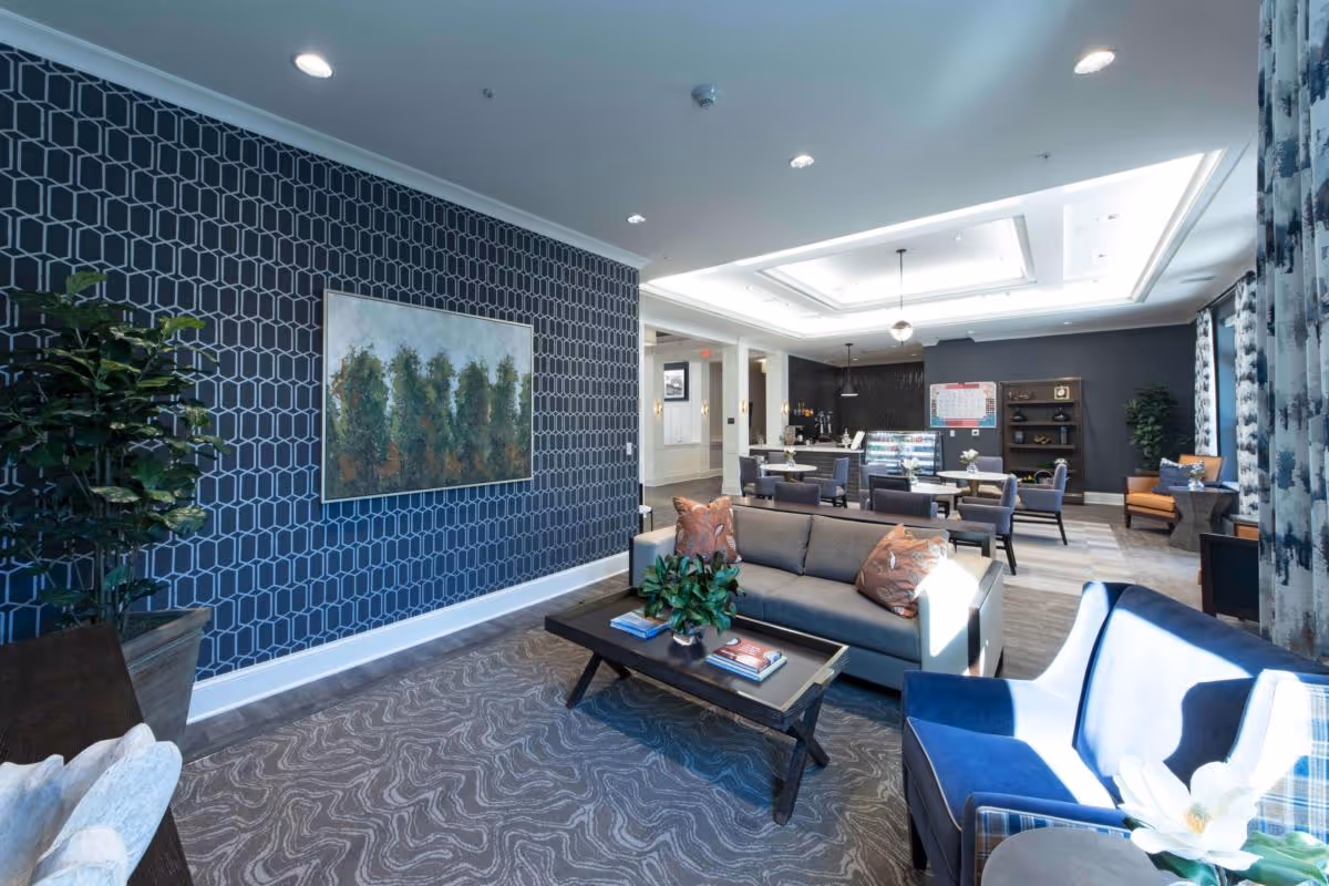 A modern and well-lit common area in a senior living facility featuring a gray sofa with decorative pillows, a dark wooden coffee table with books and a plant, a blue armchair, patterned carpet, and a wall with geometric wallpaper and a framed painting of trees. In the background, there are several tables and chairs, a counter with a display case, and additional seating with orange chairs near large windows with patterned curtains.