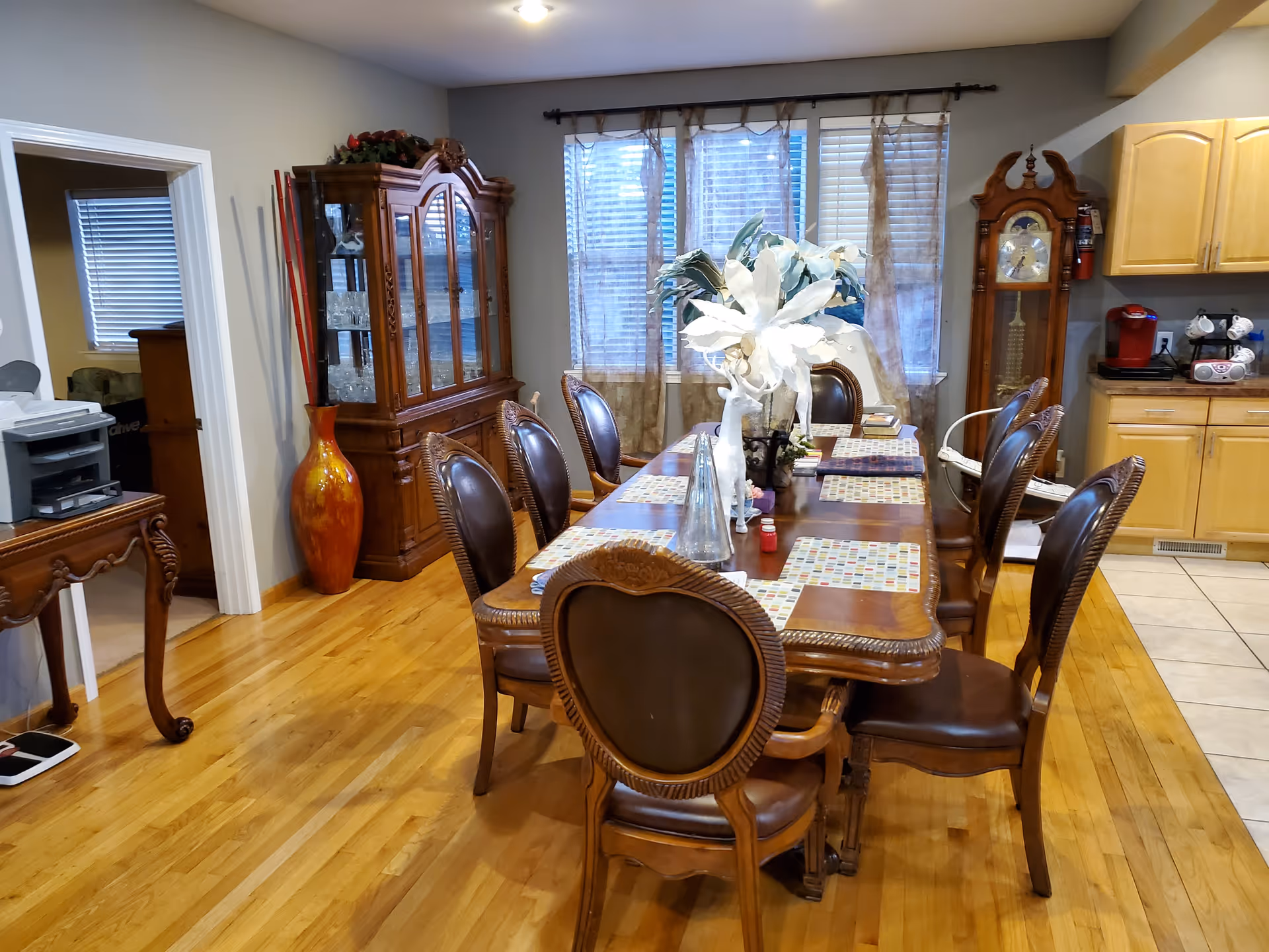A dining room with a long wooden dining table surrounded by eight ornate wooden chairs with leather seats. The table is set with placemats and decorative vases with white flowers. Behind the table, there is a large window with sheer curtains. To the left, there is a wooden china cabinet with glass doors and a tall decorative vase with red sticks. To the right, part of a kitchen with light wood cabinets and a coffee maker is visible. The floor is hardwood with a tiled section near the kitchen.