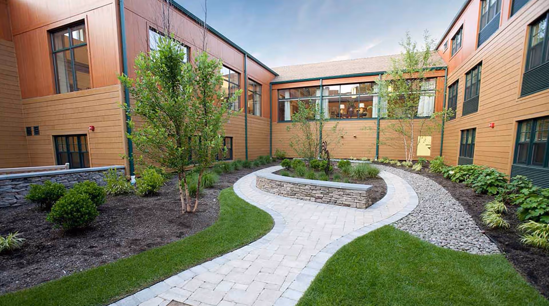Paved winding courtyard with a raised stone planter, lawn, small trees and shrubs surrounded by a multi-story wood-paneled building.