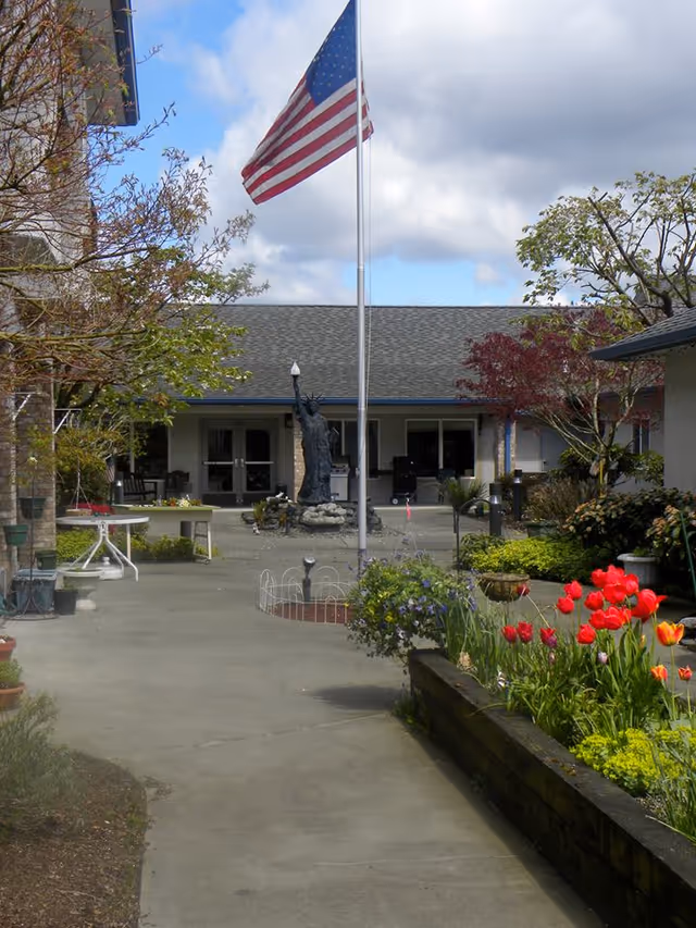 Courtyard in front of a single-story care center with an American flag on a pole, a small statue near the entrance, and flower beds with red tulips.