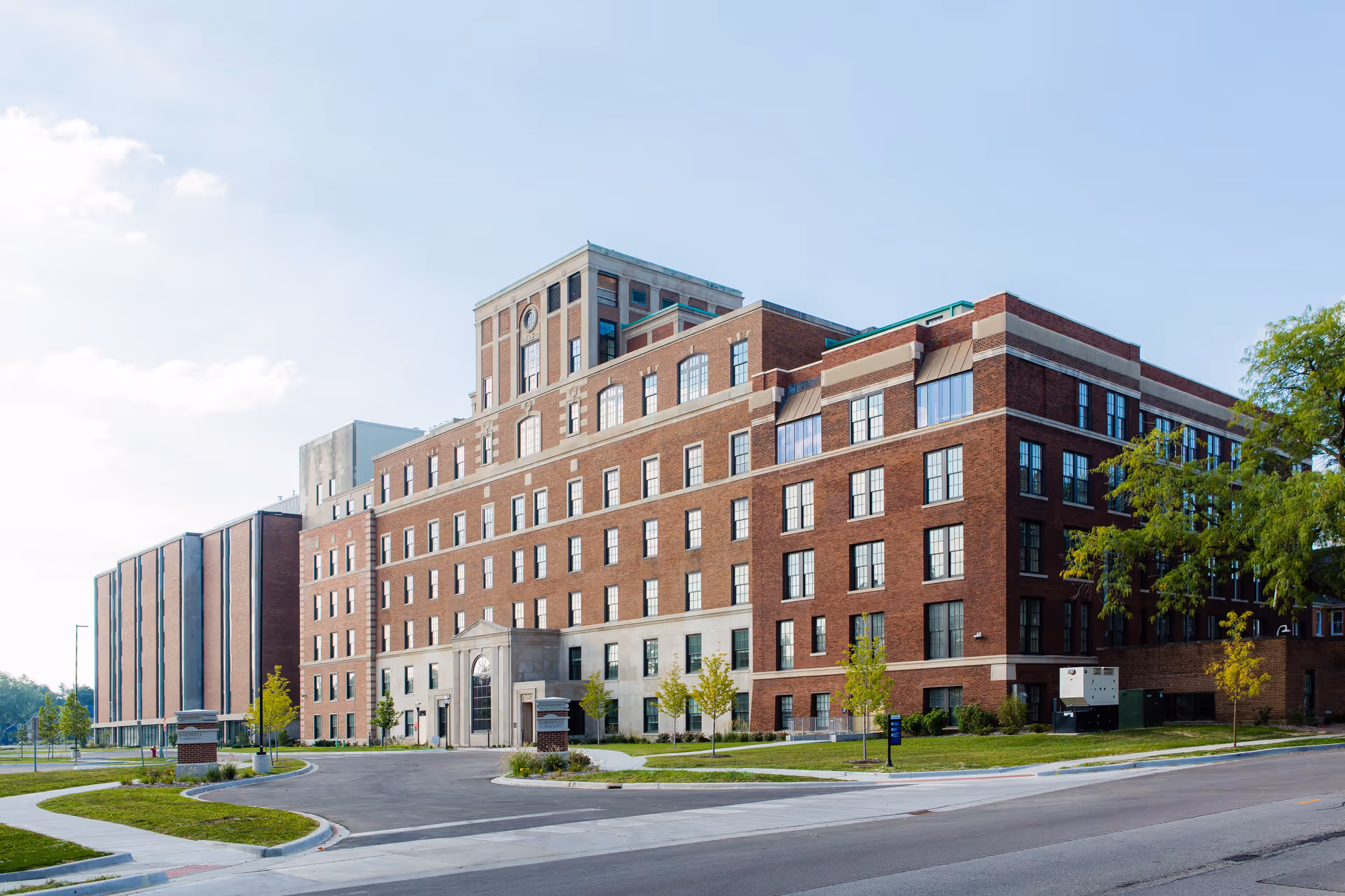 Exterior view of a large multi-story brick building with many windows, a main entrance with a stone facade, and a driveway leading up to it. The building is surrounded by small trees and grass, under a clear blue sky.