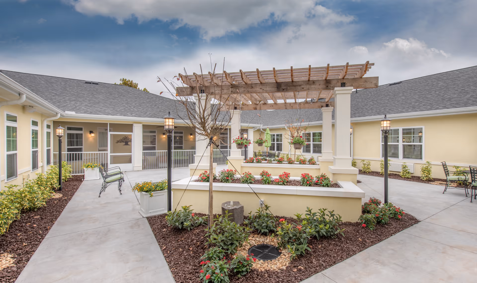 Outdoor courtyard area at The Addison of Port Orange featuring a central raised garden bed with flowers and a wooden pergola overhead. The courtyard is surrounded by a yellow building with multiple windows and outdoor seating including benches and tables with chairs. There are lamp posts lining the concrete walkways and landscaped plants around the garden beds.