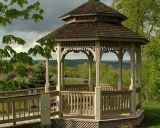 A wooden gazebo with a shingled roof situated outdoors surrounded by green trees and grass, with a cloudy sky in the background.