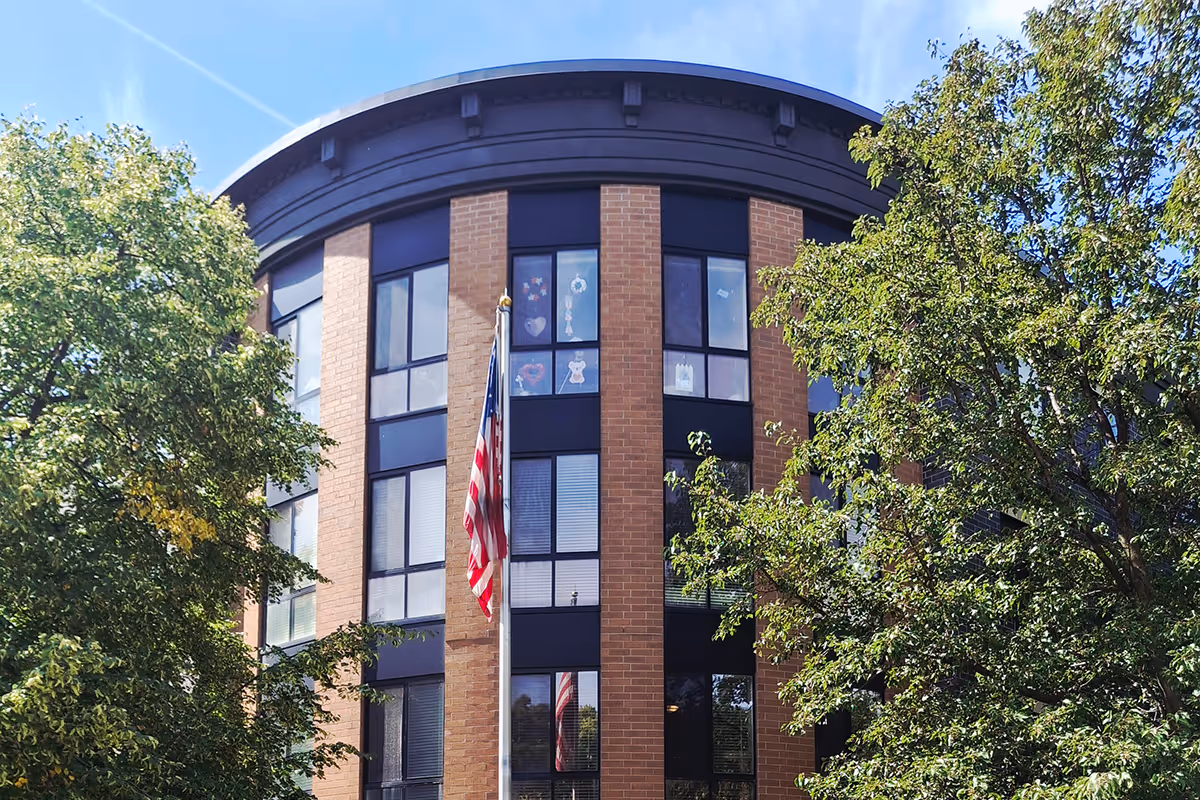 Exterior view of a brick senior living building with large windows, a flagpole with an American flag, and surrounding trees.