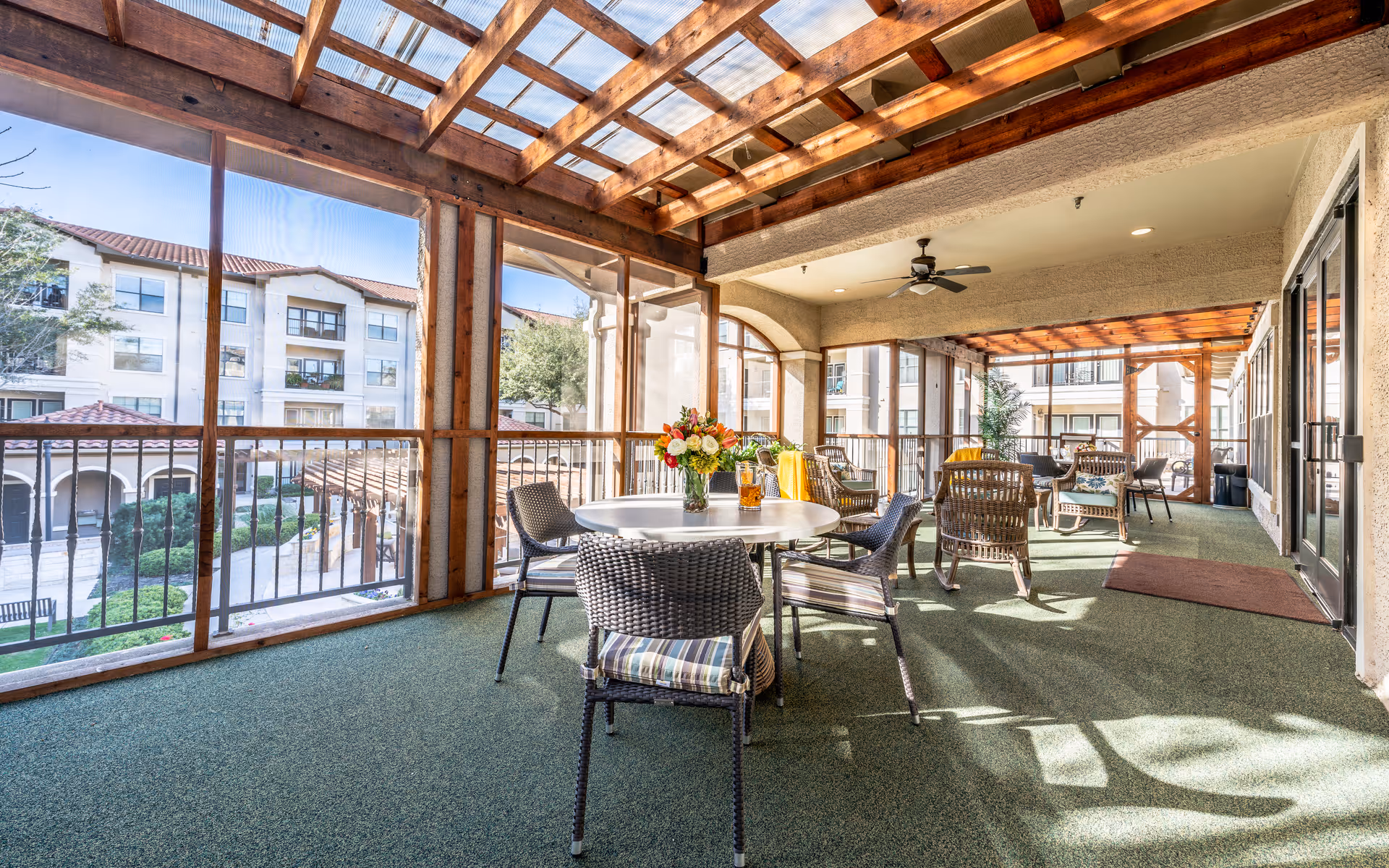 A spacious covered outdoor patio area with a wooden pergola roof and green carpet flooring. The patio is furnished with several wicker chairs and round tables, one of which has a vase with colorful flowers. The area overlooks a courtyard with a multi-story residential building in the background.