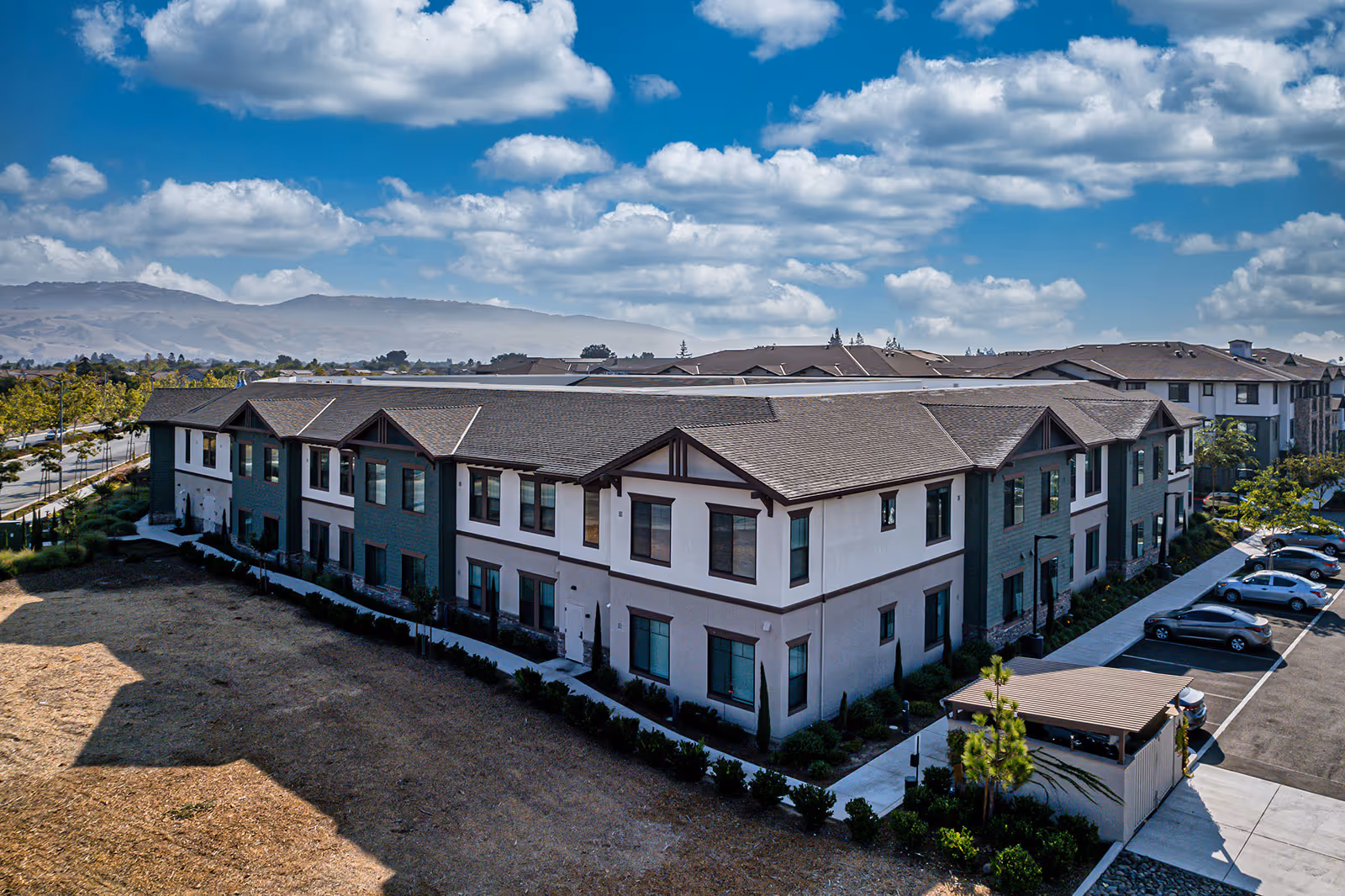 Aerial view of a large two-story senior living facility building with a brown roof and white and green exterior walls. The building is surrounded by landscaped greenery, a parking lot with several cars, and a small covered structure. The sky is blue with scattered clouds, and mountains are visible in the background.
