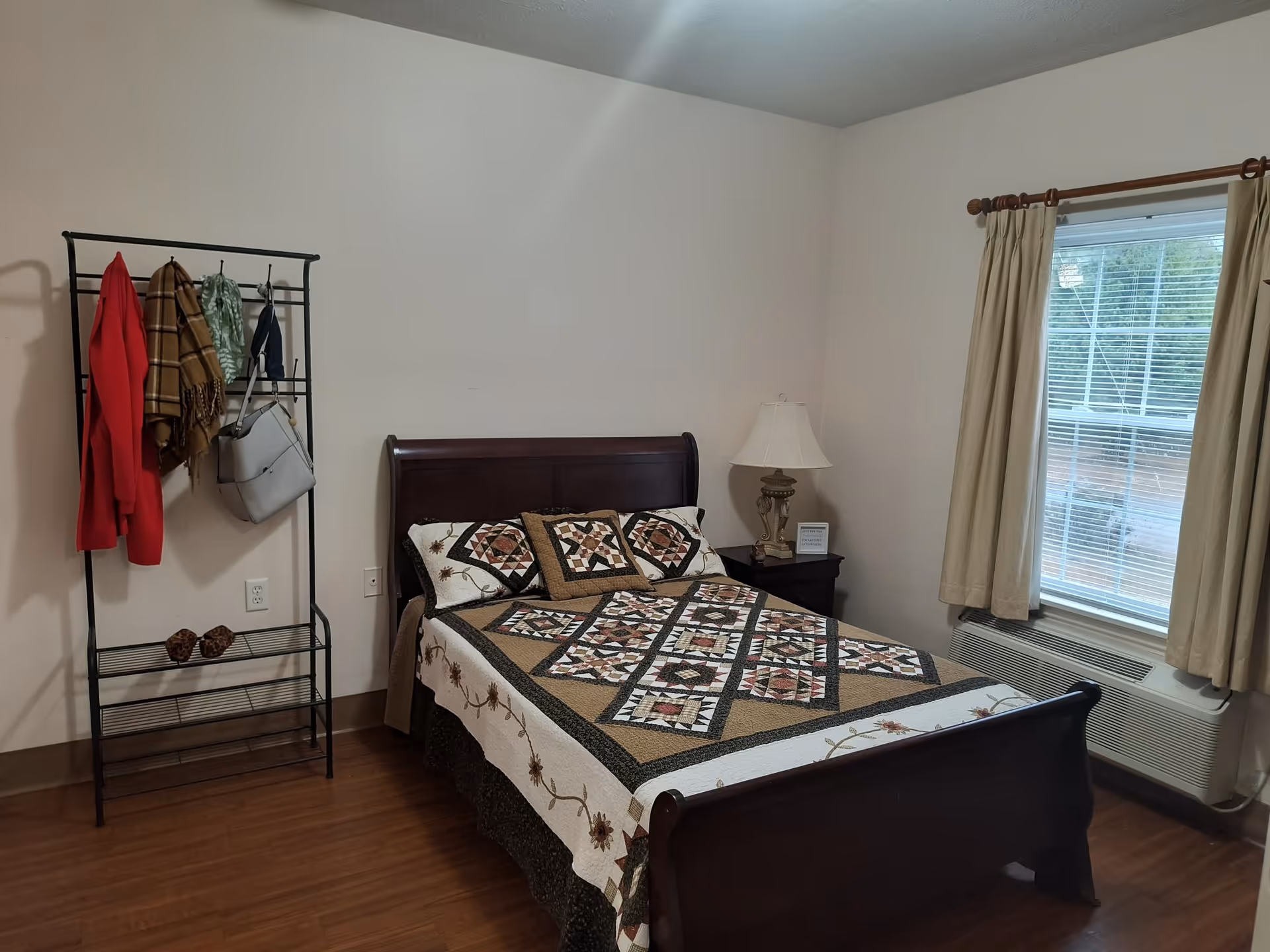 A bedroom with a wooden bed frame and a quilted bedspread featuring a geometric pattern. There is a small nightstand with a decorative lamp and a small framed item next to the bed. A window with beige curtains is on the right wall, and a black metal coat rack with several hanging items including a red coat, scarves, and a handbag is on the left side. The floor is wooden, and the walls are painted light beige.