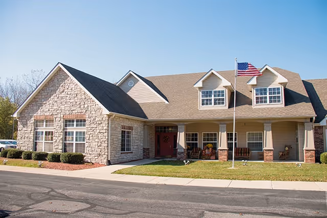 Front exterior of a stone-and-siding senior living building with a covered porch, seating, and an American flag.