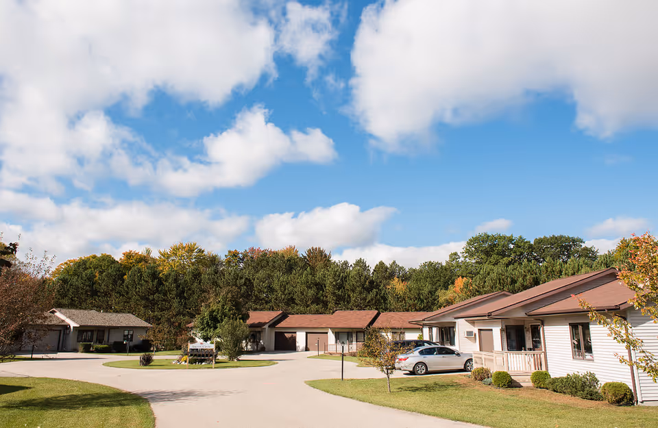Single-story senior living cottages around a paved driveway with trees and a blue sky.