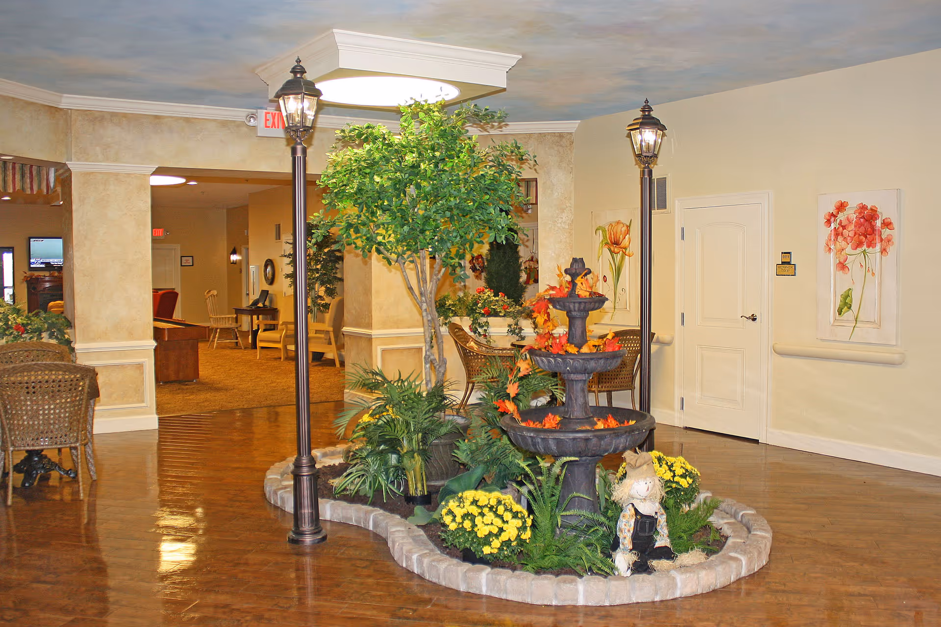 Indoor common area with a decorative garden featuring a tiered fountain adorned with autumn leaves, surrounded by green plants and yellow flowers. Two vintage-style street lamps flank the garden. Wicker chairs and tables are placed around the space, with beige walls decorated with floral paintings. The floor is polished wood, and there is an exit sign visible in the background.