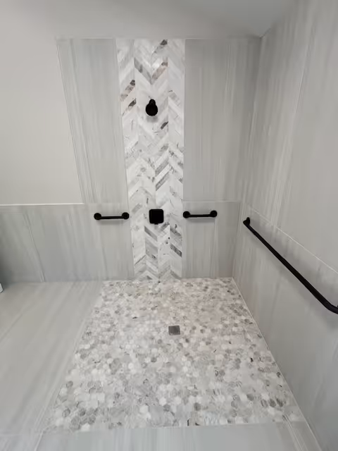 A modern walk-in shower with light gray and white tiles. The shower floor has small round mosaic tiles, and the wall features a vertical strip of chevron-patterned tiles in the center. There are three black grab bars mounted on the walls and a black showerhead and control knob.