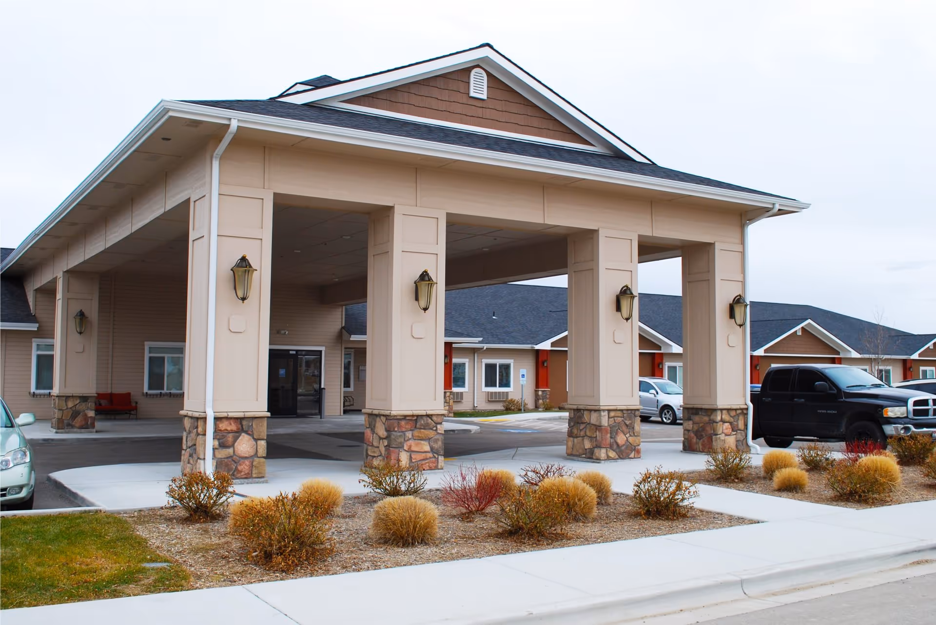 Exterior view of Aspen Creek Senior Living facility showing the covered entrance with stone pillars, parked cars, and landscaped bushes in front.