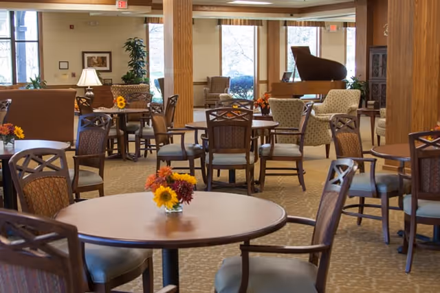 A spacious and well-lit common area with multiple round wooden tables and chairs arranged throughout the room. Each table has a small floral centerpiece. The room features large windows letting in natural light, comfortable armchairs, wooden columns, and a grand piano in the background.