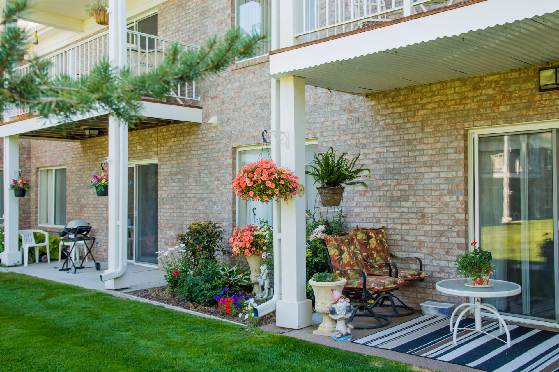 Outdoor patio area of a brick building with hanging flower baskets, potted plants, a cushioned bench, a small round table with a plant, and a striped outdoor rug. There is green grass in the foreground and sliding glass doors leading inside.