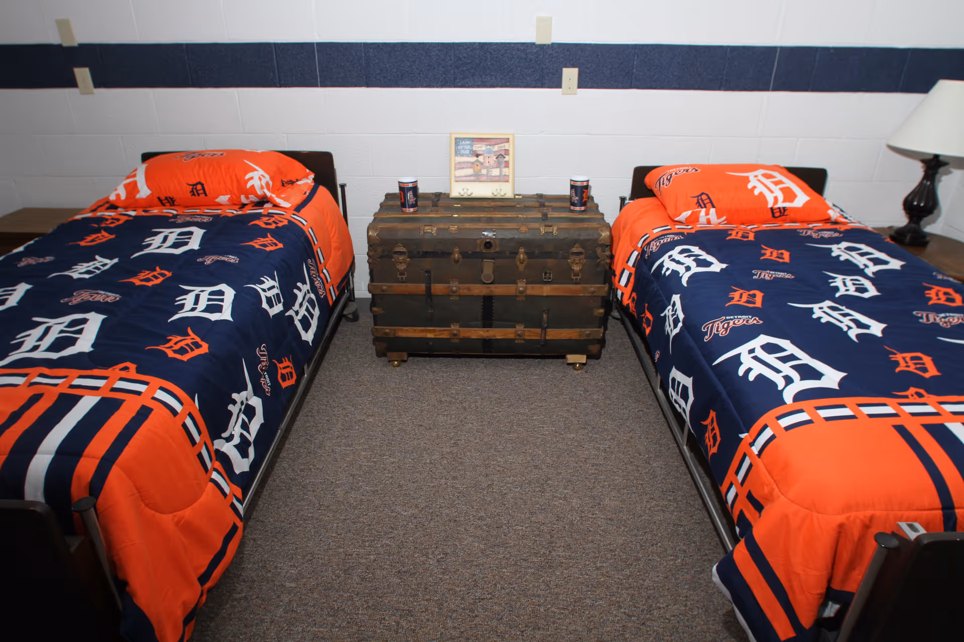Two twin beds with Detroit Tigers blue and orange bedding separated by a vintage trunk in a small bedroom.