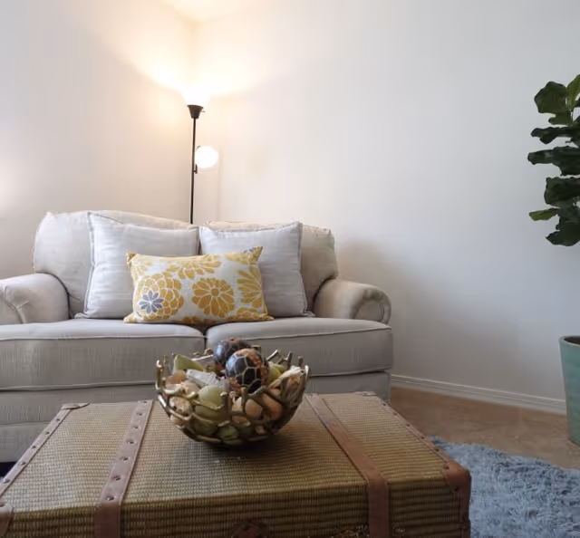 A cozy living room area with a beige sofa adorned with three cushions, including one with a yellow floral pattern. In front of the sofa is a woven trunk-style coffee table with a decorative bowl containing assorted natural elements. A tall floor lamp with two lights is positioned behind the sofa, and a large potted plant is visible on the right side against a plain white wall.