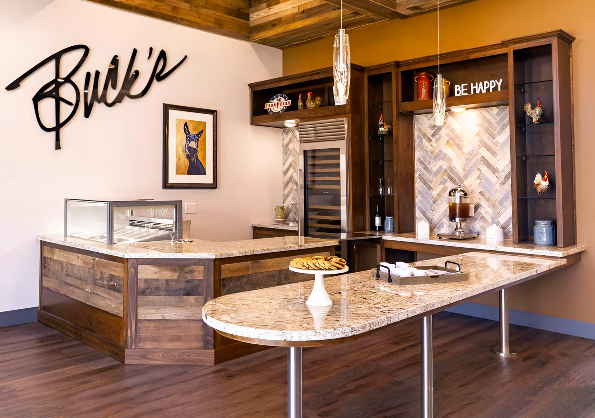 A modern kitchen area with wooden cabinets, a granite countertop island with cookies on a white cake stand, a beverage dispenser, and decorative items including a framed picture of a donkey and a sign that says 'BE HAPPY'. The wall has a wooden sign that reads 'Buck's'.