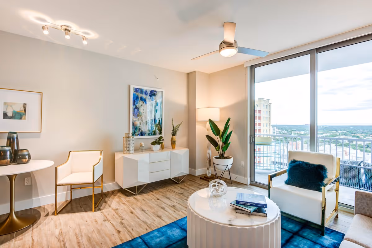 Bright and modern living room with light wood flooring, a white round coffee table with books and decorative items, a white armchair with gold accents and a dark blue cushion, a white sideboard with plants and decor, a floor lamp, and large sliding glass doors leading to a balcony with a city view.