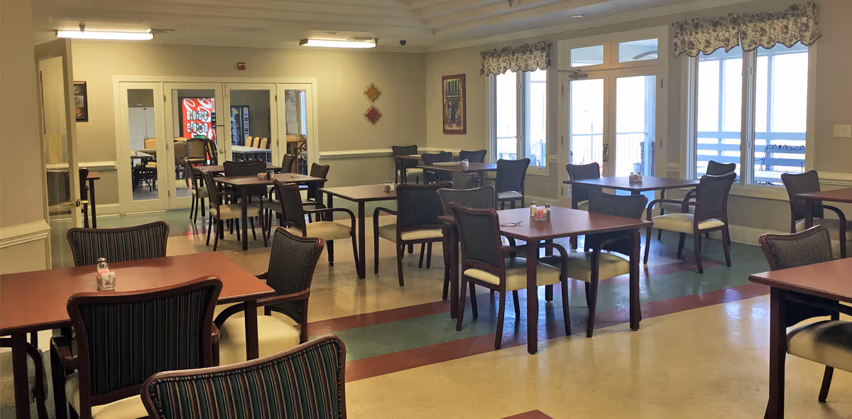 Spacious dining room with several square wooden tables and cushioned chairs arranged near large windows and glass doors.