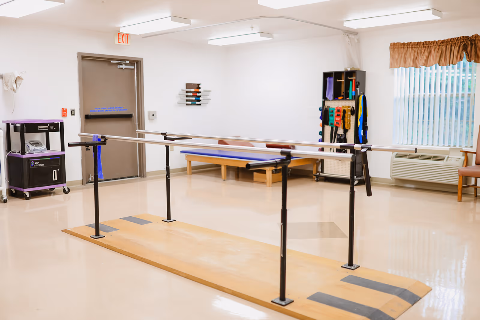 A rehabilitation room with parallel bars for walking exercises on a wooden platform, a blue padded therapy table, exercise equipment on a rack, a window with vertical blinds and a brown valance, an air conditioning unit below the window, and a closed exit door.