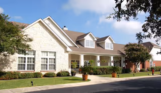 Exterior view of a single-story senior living facility building with a stone facade, multiple windows, a covered porch with hanging plants, and a well-maintained lawn and landscaping under a partly cloudy sky.