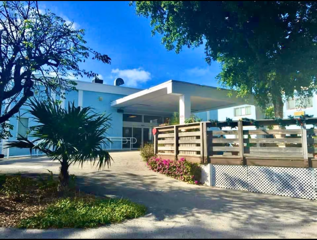 Exterior view of Oasis at the Keys Nursing & Rehab facility showing the entrance with a covered walkway, surrounded by trees, plants, and a wooden fence with flowers along the pathway.