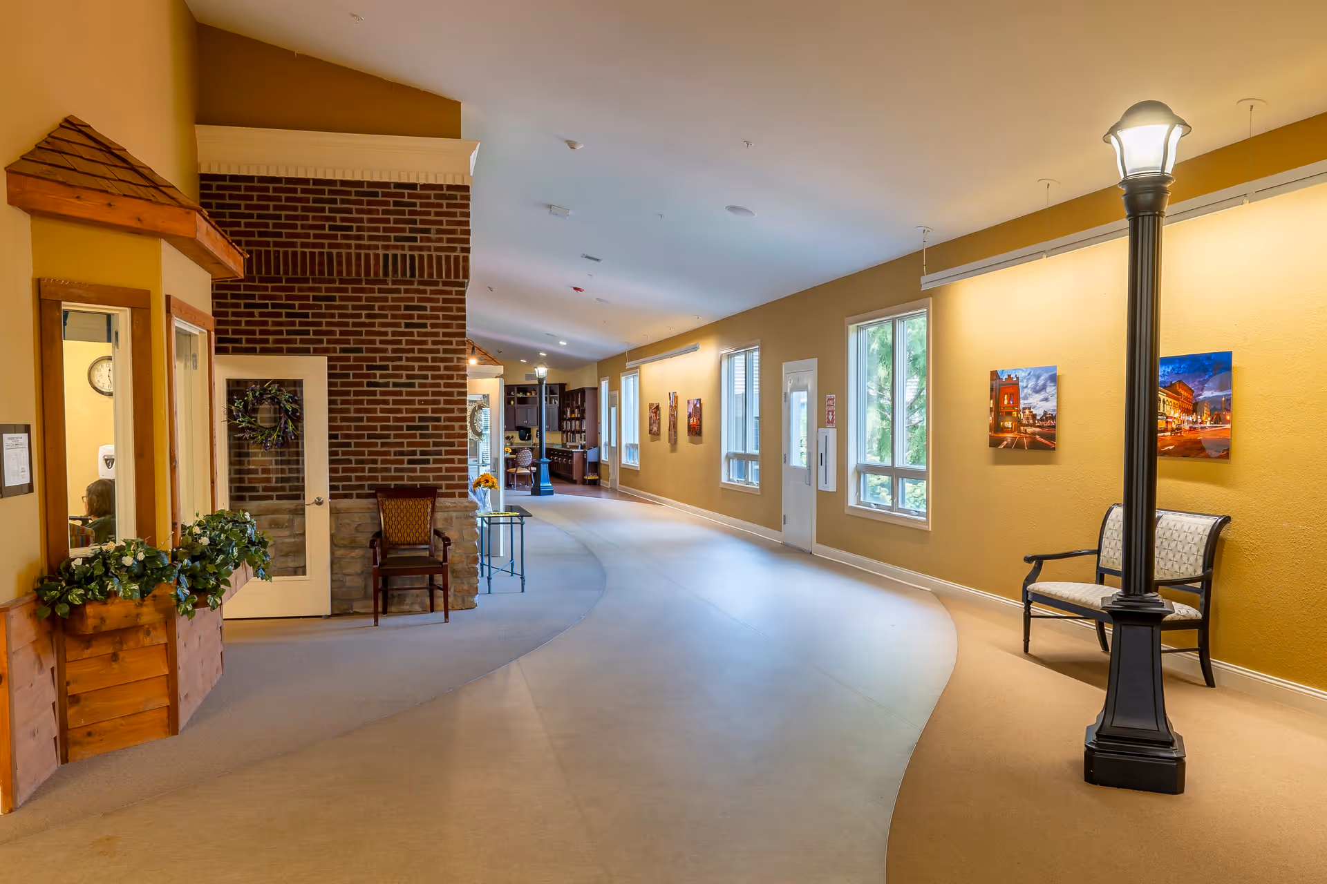 A wide, well-lit hallway in a senior living facility with beige walls and carpeted floor. The hallway features a brick accent wall with a door and chair, several windows letting in natural light, framed pictures on the walls, a decorative streetlamp-style light fixture, and seating benches along the walls.