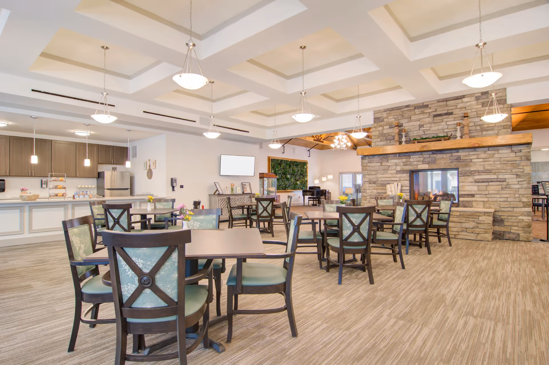 A spacious dining area in a senior living facility with multiple tables and chairs arranged neatly. The room features a stone fireplace, pendant lighting, and a kitchen area with cabinets and a refrigerator in the background. There is also a piano and decorative greenery on the walls.