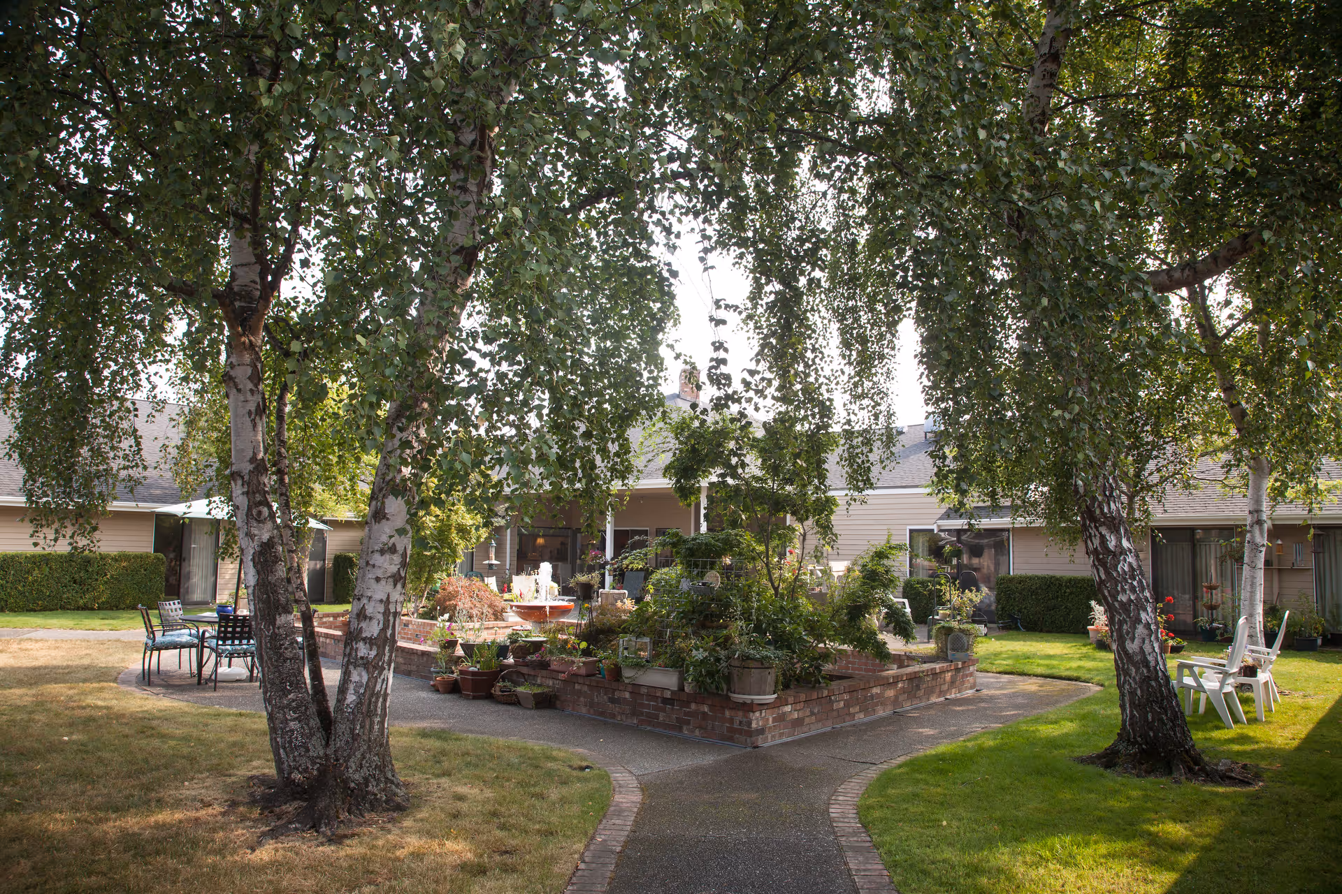 Outdoor courtyard area at Cap Sante Court Retirement Community featuring a paved walkway, green grass, several trees, a raised brick planter with various plants, and outdoor seating including tables and chairs.