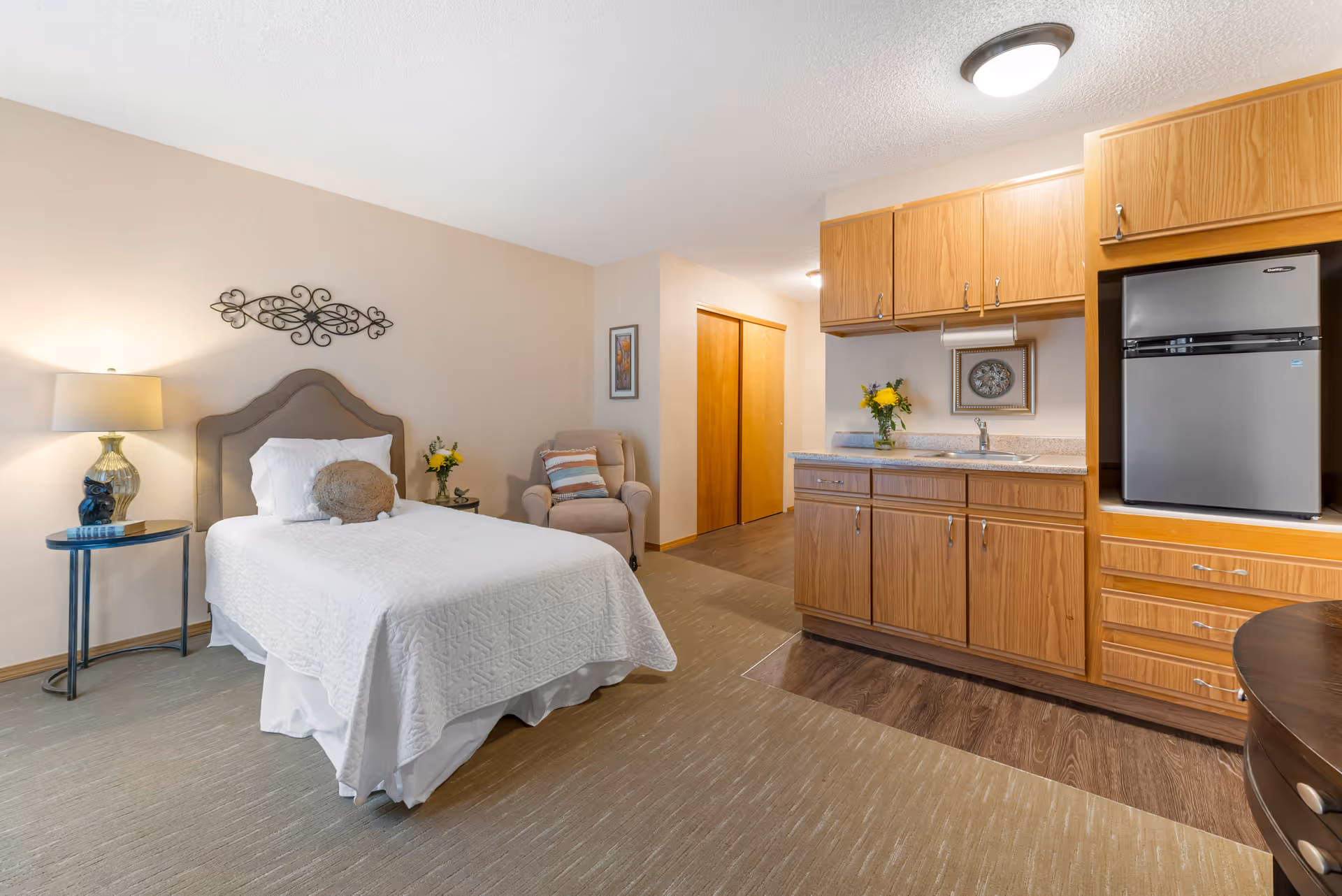 A studio-style room with a single bed, bedside table and lamp, armchair, and a small kitchenette with wood cabinets and a refrigerator.