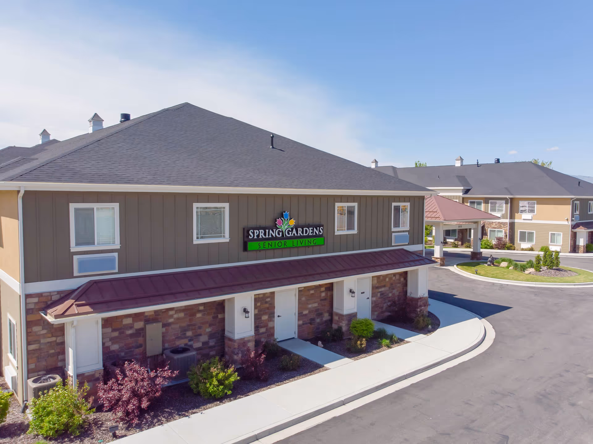 Exterior view of the Spring Gardens Senior Living facility showing a two-story building with a dark roof, beige and brown walls, and a sign that reads 'Spring Gardens Senior Living'. The building is surrounded by a paved driveway and landscaped with bushes and small trees under a clear blue sky.