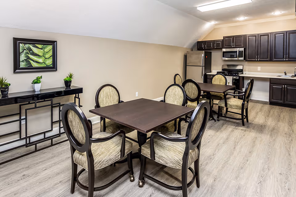 A communal dining area with square tables and upholstered chairs and a kitchenette with stainless appliances and dark cabinets in the background.
