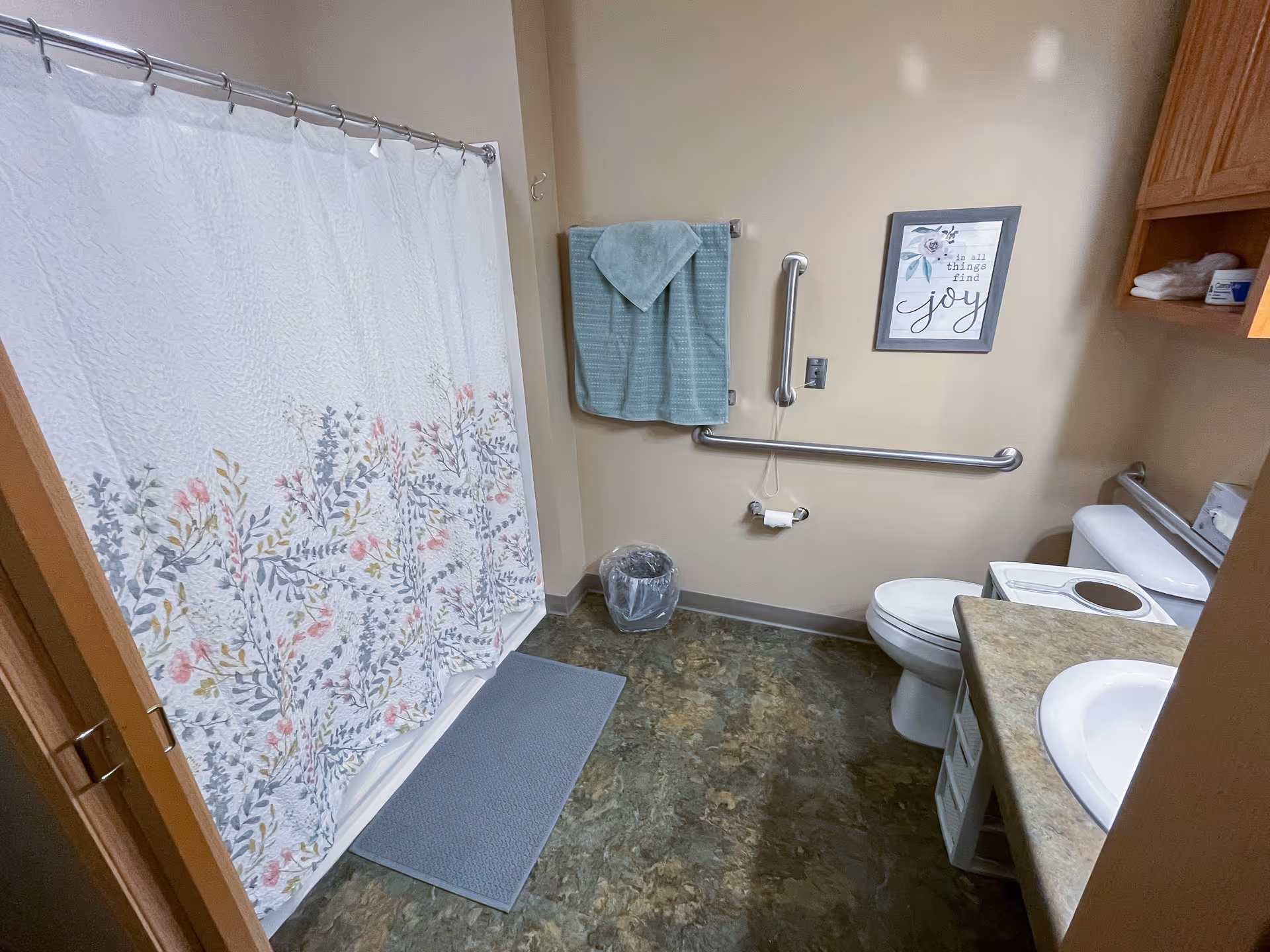 A bathroom with a floral-patterned shower curtain, a gray bath mat, a towel rack with a folded green towel, a trash bin, a toilet with grab bars on the wall, a countertop with a sink, and a framed wall decoration that reads 'in all things find joy'.