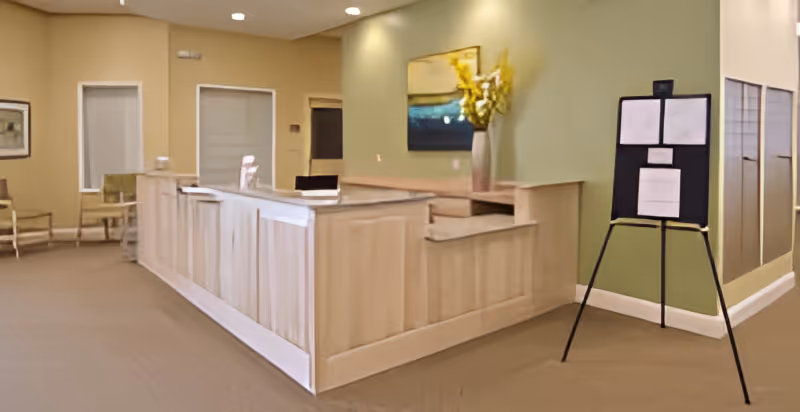 Reception area with a light wood front desk, a vase with yellow flowers on the counter, a painting on the green wall behind the desk, a black stand with papers on the right, and chairs along the beige wall on the left.