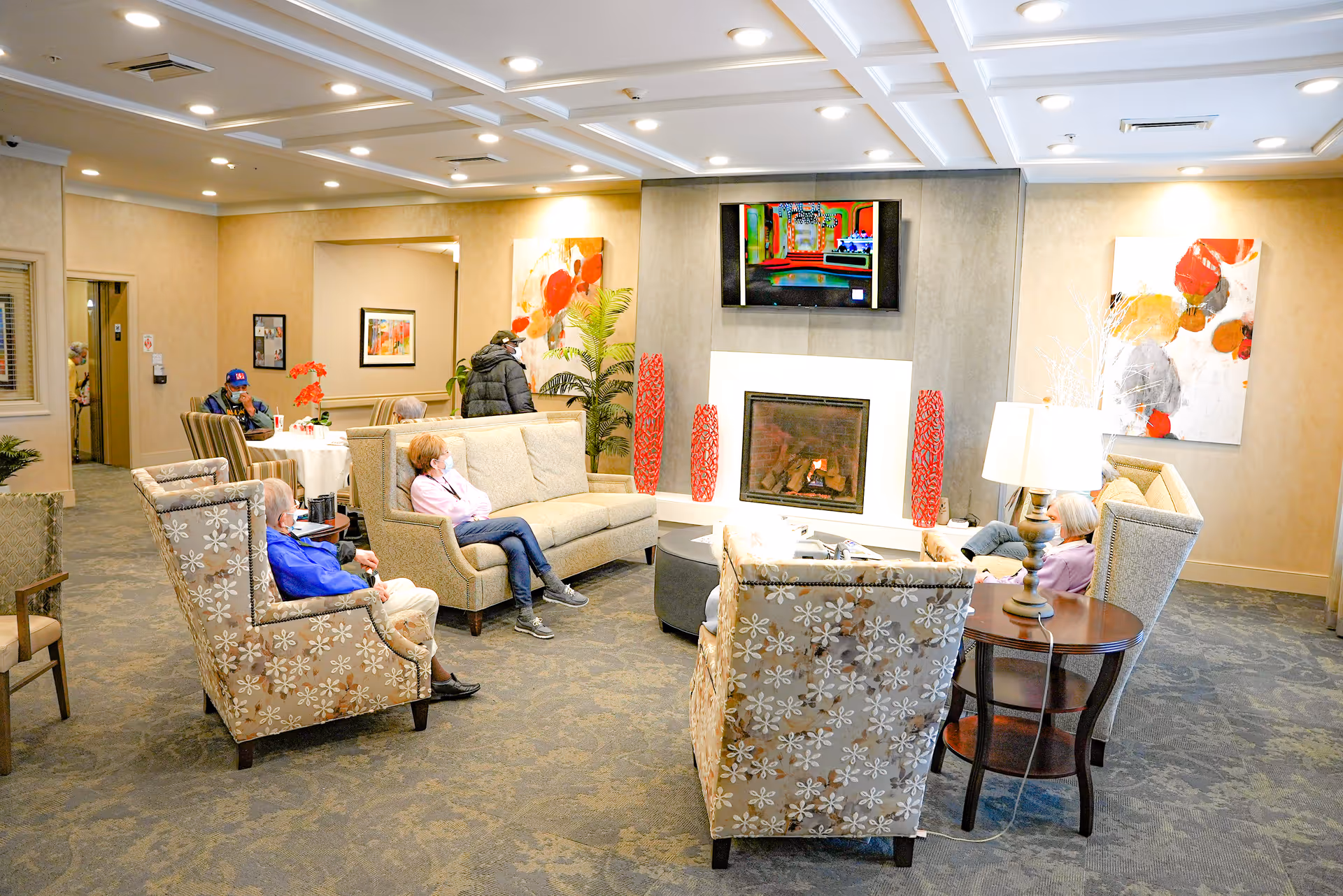 A cozy senior living facility common area with several elderly people seated on patterned armchairs and sofas around a fireplace. A flat-screen TV is mounted above the fireplace, and the room is decorated with modern abstract paintings and plants. The ceiling has recessed lighting, and the carpet has a subtle pattern.