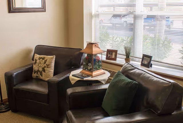 A cozy seating area with two dark leather armchairs, one with a floral cushion and the other with a green cushion, separated by a small table holding a decorative lantern. Behind the chairs is a window with blinds, a small potted plant, and framed photos on the windowsill.