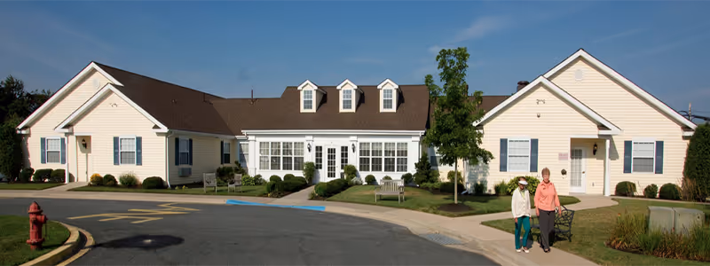 Front view of a single-story pale yellow assisted living building with a central glass entry, landscaped lawns, and two people walking on the sidewalk.