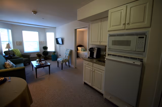 Interior view of a senior living facility apartment showing a living room with a green armchair, a coffee table, a patterned chair, and a wall-mounted TV. To the right, there is a kitchenette area with white cabinets, a microwave, a small refrigerator, and a coffee maker. A bedroom with a bed and pillows is visible through an open doorway.