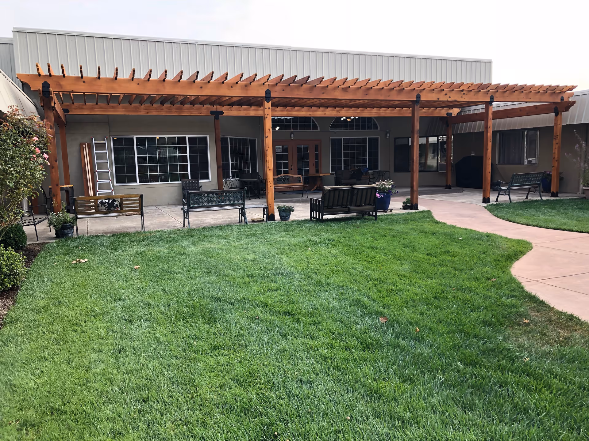Outdoor patio area at Grace Manor Senior Living with a wooden pergola covering several benches and chairs. There is a well-maintained green lawn in the foreground and a paved walkway curving through the grass. The building exterior features large windows and doors behind the seating area.